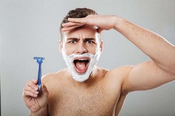 Expressions faciales amusantes d'un homme mal rasé effectuant une procédure matinale avec un rasoir et de la crème sur le visage sur un mur gris.
