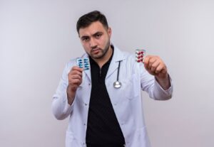 Jeune médecin barbu en blouse blanche avec stéthoscope, tenant des ampoules contenant des pilules et les montrant à la caméra d'un air sérieux.