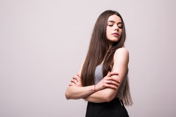 Portrait d'une jeune jolie femme élégante souriante en t-shirt gris sur un mur blanc, isolée, aspect naturel, longs cheveux bruns, bras croisés