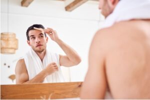 Jeune homme mal rasé se peignant les cheveux, debout, une serviette blanche sur le cou, près d'un miroir dans une salle de bains.