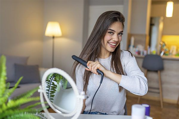 Une belle femme souriante se repasse les cheveux et autour d'elle des détails à utiliser. , La décoloration des cheveux est mauvaise pour la santé 