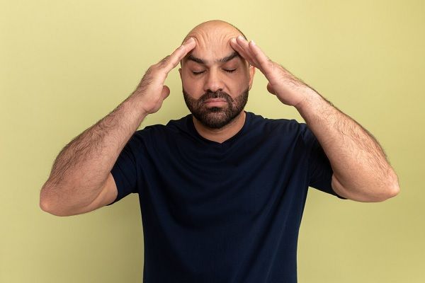 Homme barbu en t-shirt noir touchant sa tête ayant l'air malade souffrant de maux de tête debout sur un mur vert La santé mentale affecte les cheveux.