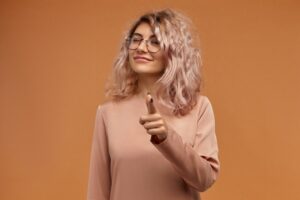 Je te regarde. Portrait horizontal d'une jeune femme caucasienne joyeuse et enjouée, portant des lunettes rondes et un anneau dans le nez, pointant son index vers l'appareil photo et faisant un clin d'œil. Langage corporel
